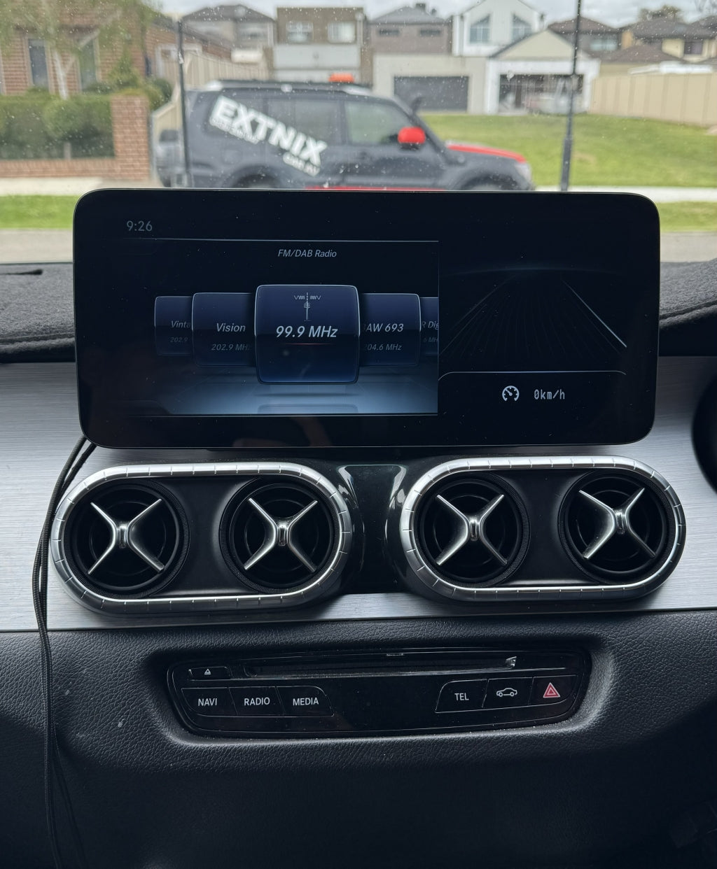 Merc Xclass w205 dashboard with a navigation screen displaying a radio station, with a vehicle and house in the background.